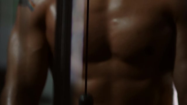 Mid-section of muscular shirtless athlete with tattooed arm doing cable rope triceps pushdowns in gym