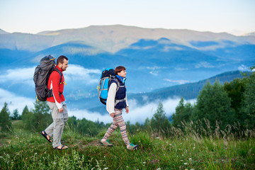 Walking in the mountains. The guy and the girl with backpacks are on the path on a hilltop with a beautiful landscape of mountains in the haze. Concept of active outdoor hiking lifestyle