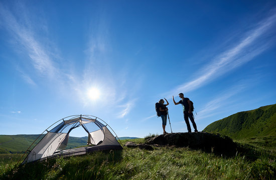 Two young people in backpacks and trekking sticks give each other a high five near the camping in the Carpathian mountains under the blue sky with a bright sun.