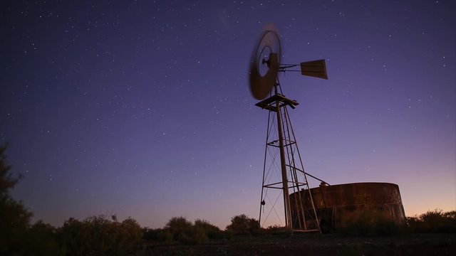 Long wide shot time lapse from dusk to night stars of a wind pump or wind mill rotating in the wind to generate renewable electricity power with land and trees.