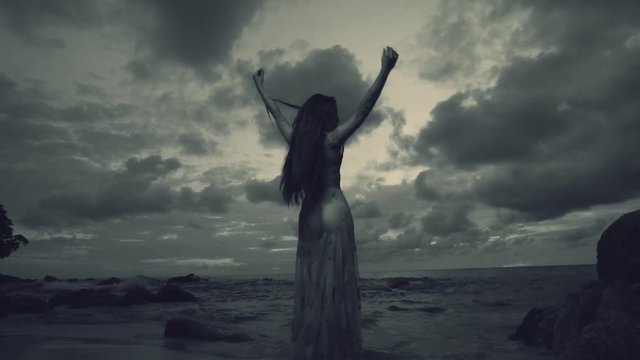 Back view of beautiful mysterious woman in long dress at the sandy beach near rocks over sea and cloudy sunset sky background - black and white video in slow motion