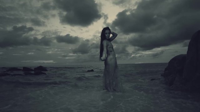 Beautiful mysterious woman in long dress at the sandy beach near rocks over sea and cloudy sunset sky background - black and white video in slow motion
