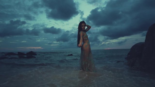 Beautiful mysterious woman in long dress at the sandy beach near rocks over sea and cloudy sunset sky background - video in slow motion