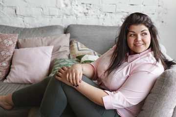 Horizontal portrait of charming body positive plus size adorable young woman sitting on sofa with her legs bent, dressed in black jeans and pink shirt, feeling carefree while relaxing at home