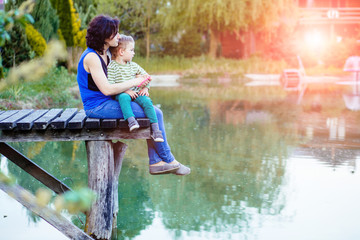 Side view mother and baby son sit togather on the wooded bridge on lake in summer evening time. Family, summer, holiday, love, motherhood concept.