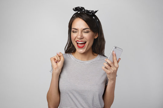 Picture of cheerful overjoyed young woman winner having happy excited look, screaming Yes and raising clenched fist, being lucky to win electronic gadget in lottery, posing in studio with cell phone
