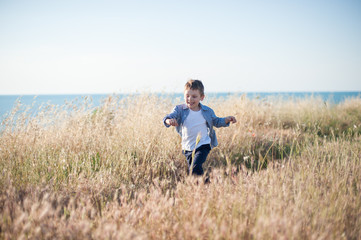 Naklejka na ściany i meble happy caucasian smiling little kid running among yellow grass on sea background in summer sunny day
