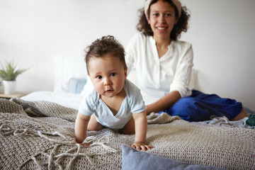 Healthy active infant with chubby cheeks and big brown eyes having curious look, staring at camera, making discoveries while crawling along parents' bed, his mom smiling broadly in background