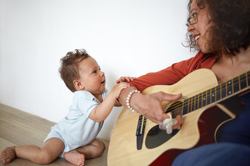 Happy young Hispanic female musician sitting on floor, playing guitar to her adorable cute baby son...