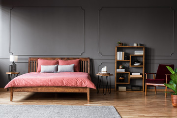 Real photo of a spacious bedroom interior with pink bed standing against black wall with molding next to a creative bookshelf and red armchair