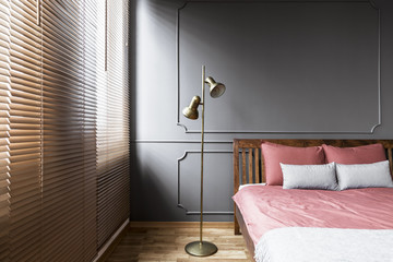 Blinds and golden lamp in dark and elegant bedroom interior with pink sheets on a wooden bed and molding on the wall. Real photo