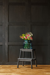 Fototapeta na wymiar A rich colorful bouquet of beautiful summer flowers in a blue glass vase on a black step stool in a dark living room interior