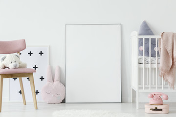Plush toy on pink wooden chair next to empty poster with mockup in baby's room interior. Real photo