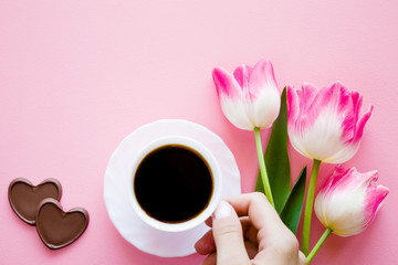 Woman's hand holding white cup of black coffee. Tulips on pastel pink table. Resting and enjoying with coffee and sweets. Dating and love. Empty place for inspirational, motivational text or quote.