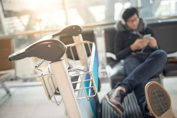 Fototapeta premium Young asian man using smartphone and listening to music while waiting for connecting flight on bench in the international airport terminal, travel abroad concept