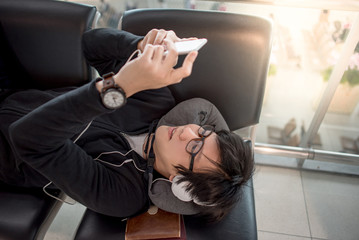 Fototapeta na wymiar Young Asian man with eyeglasses and headphones using smartphone and listening to music on bench while waiting for connecting flight in the international airport terminal, travel lifestyle