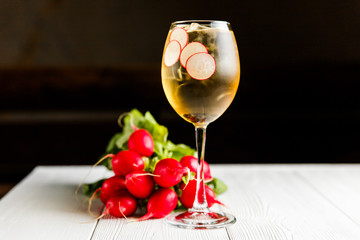 A photo of a light yellow cold summer aperitif alcoholic cocktail in a wine glass, full of ice cubes, garnished with radish. White wooden table.