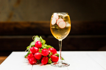 A photo of a light yellow cold summer aperitif alcoholic cocktail in a wine glass, full of ice cubes, garnished with radish. White wooden table.