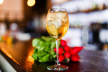 A photo of a light yellow cold summer aperitif alcoholic cocktail in a wine glass, full of ice cubes, garnished with radish. White wooden table.