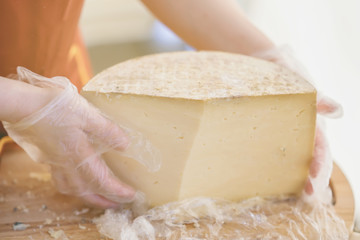 Half of a large head of cheese in the hands of the seller in gloves, wooden board. Selective focus. Gastronomic dairy produce, real scene, food market