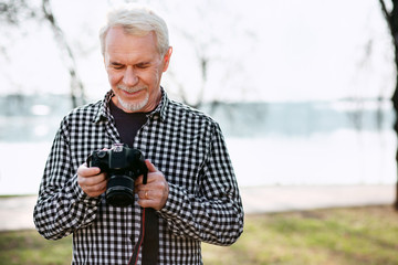 Creative hobby. Happy senior man posing on the blurred background and using camera