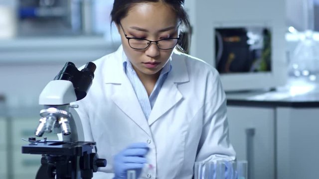 Medium shot of young Asian woman in lab coat, glasses and rubber gloves studying chemical reactions with microscope when working in laboratory