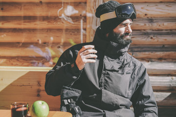 Bearded cool snowboarded taking rest after ride session.Young man drinking cup of hot tea on sunny terrace. Blurred background.Horizontal.Visual effects - Powered by Adobe