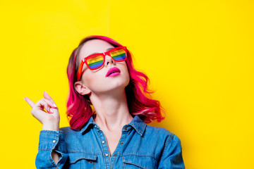 Young pink hair girl in blue shirt and rainbow glasses. Portrait isolated on yellow background