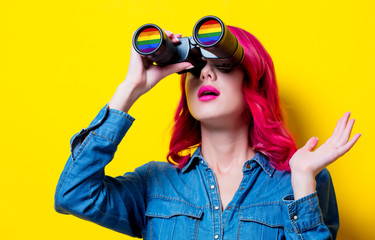 Young pink hair girl in blue shirt holding a binoculars with rainbow. Portrait on isolated yellow...