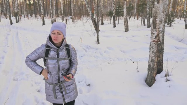 Beautiful woman is walking in the woods. Traveler is walk in birch forest city park. Girl walks in evening time with beautiful juicy sunset. Explorer is dressed in stylish silver down jacket and