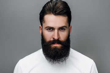 Fototapeta premium Horizontal close up portrait of handsome brutal masculine young bearded male looking at the camera, posing on a gray studio background. Portrait of young European fashion barber with trendy beard.