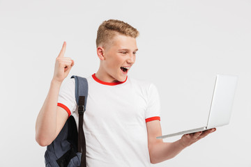 Image of young happy guy 16-18 years old wearing t-shirt and backpack pointing finger up at copyspace while holding laptop, isolated over white background
