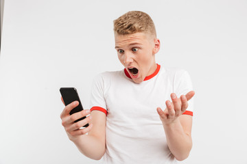 Photo of outraged young man wearing casual style screaming while holding and using smartphone, isolated over white background