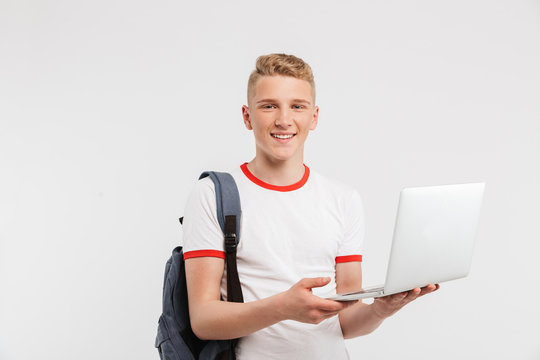 Image of teen man 16-18 years old wearing basic clothing and backpack holding open laptop with beautiful smile, isolated over white background