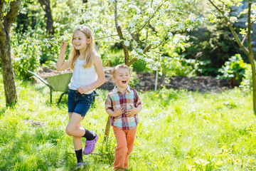 Emotional positive shot of two cute little gardeners sister and brother over wheelbarrow on background in yard or garden at bright sunny summer day. Copy space text