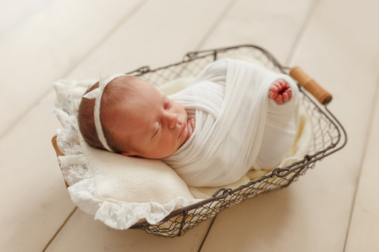 Cute sleeping newborn girl with a bandage on her head on a small pillow, wrapped in a white lump, in a metal basket on a wooden floor