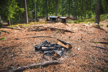 Campfire place on a top of a hill, on the ground covered by fallen pine needles. Two wooden shelters surrounded by pines in the blurry background. Taken in nature reserve Ånnaboda in central Sweden.