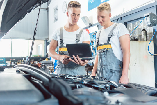 Experienced auto mechanic using a laptop for scanning and interpreting engine error codes next to an apprentice in a modern automobile repair shop