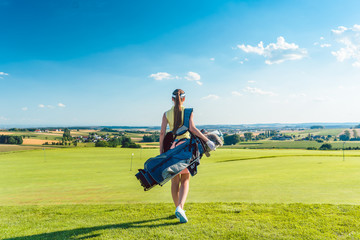 Full length rear view of an active woman carrying a blue stand bag, while walking on the grass towards a training area for professional golf in a sunny day