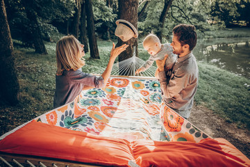 happy hipster family playing with cute little son on hammock in summer sunny park. stylish mom and dad with kid relaxing in forest and smiling, having fun. space for text. family vacation