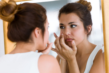 Beautiful young woman removing pimple from her face in a bathroom home.