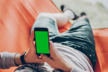 hipster man holding smart phone with empty screen with space for text, and relaxing in hammock in sunny summer park. mock up. guy looking at blank phone, social media concept