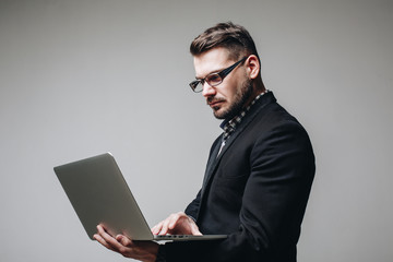 Side view shot of handsome business man in glasses and black suit holding laptop in hands and writing something. Isolated on gray background