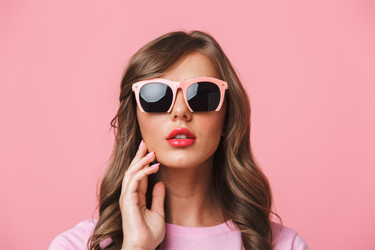 Photo closeup of sexual charming woman 20s with long curly hairstyle wearing trendy sunglasses looking at camera with fashion look, isolated over pink background