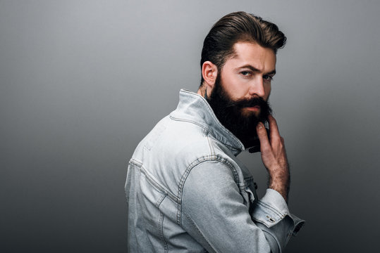 Rear view portrait of stylish brutal young Caucasian male with thick beard wearing trendy denim jacket, staring at camera with serious look. Handsome barber man posing against gray studio wall