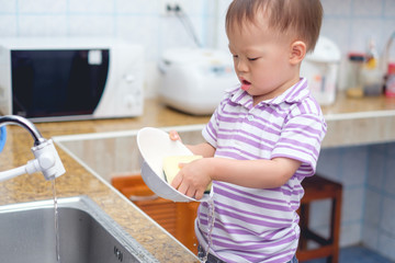 Cute little Asian 2 year old toddler boy child standing & having fun doing the dishes, concentrate on washing dishes in kitchen at home, Little home helper, chores for kids, child development concept