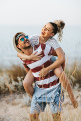 Portrait of man carrying girlfriend on his back along the sea shore. Man giving piggyback ride to...