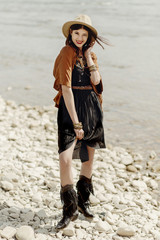 beautiful stylish boho woman smiling, with hat, fringe poncho and boots. girl in gypsy hippie look young traveler posing near river rocks in mountains. sensual look. atmospheric moment. summer travel