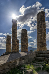 Delphi Town, Phocis - Greece. The Temple of Apollo (Olympian god of sun in ancient greek mythology) at the archaeological site of Delphi. Sun shines above the ancient columns of the temple. Cloudy sky