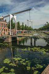Fototapeta na wymiar Bascule bridge over the canal with aquatic plants and brick houses on a sunny day in Weesp. Quiet and pleasant village full of canals and green near Amsterdam. Northern Netherlands.